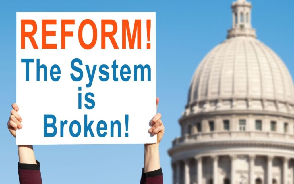 hands hold up a sign that reads reform the system is broken next to a capital building 